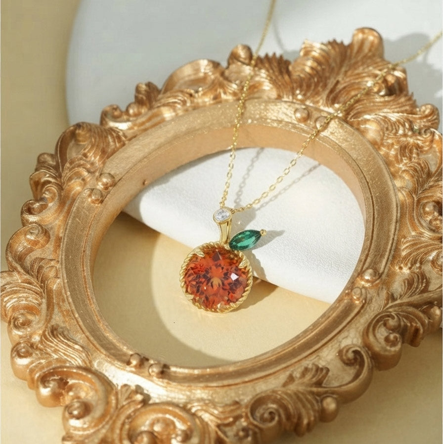 A gold pendant necklace with a round orange-red gemstone and a small green accent stone, displayed on a white stand inside an ornate gold picture frame.