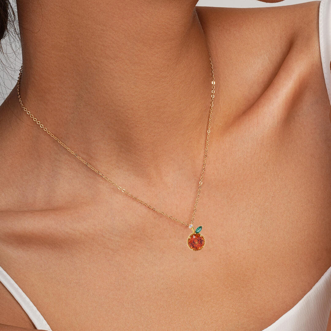 A delicate gold chain necklace with a small pendant featuring a warm orange-red stone and a tiny green accent, shown worn on a model's neck.