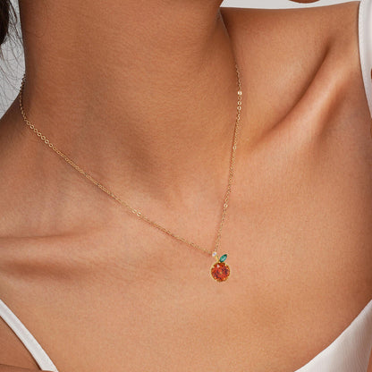 A delicate gold chain necklace with a small pendant featuring a warm orange-red stone and a tiny green accent, shown worn on a model's neck.